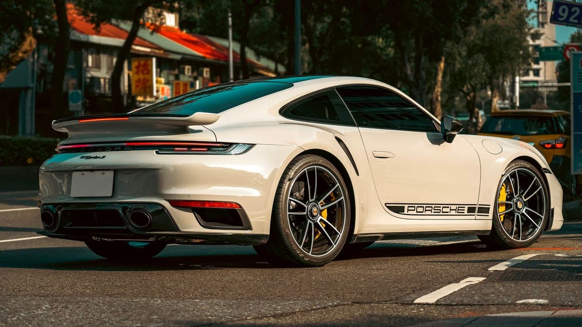 Photo: David Ford / Unsplash White Porsche 911 Turbo S coupe seen from the rear three-quarter on a sunlit city street, with yellow brake calipers, a black Porsche side stripe and a blank rear plate.