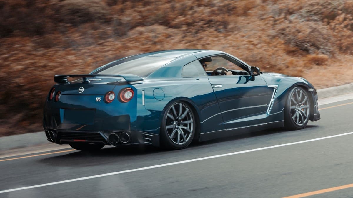 Photo: Stephan Louis / Pexels Dark blue Nissan GT-R R35 driving along a road past a brown hillside, seen from the rear three-quarter.